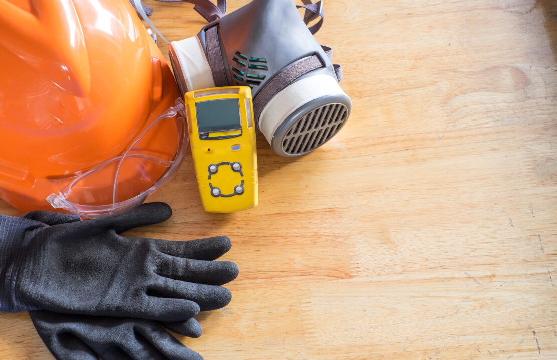 A yellow gas monitoring device leaning up against a respirator. Next to the monitoring device is an orange hard hat and a pair of black gloves.